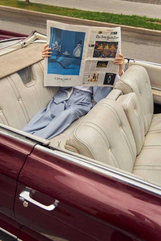 Person reading newspaper in a vintage convertible car with white leather seats and maroon exterior.