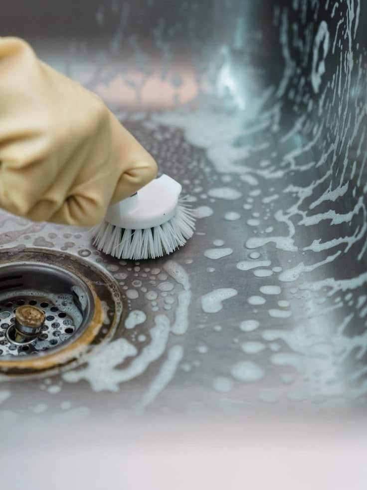 Gloved hand scrubbing soapy stainless steel sink with a brush for deep cleaning.