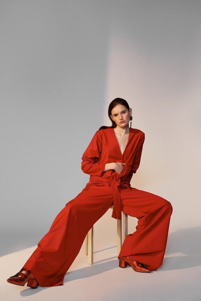 Woman in red jumpsuit sitting on stool, striking pose under soft lighting.