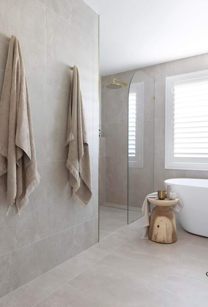 Modern bathroom with free-standing tub, glass shower, beige tiles, and wood stool for a minimalist design.
