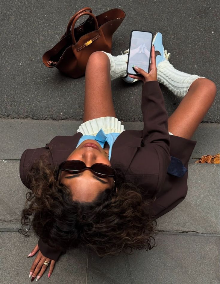 _ 47 Woman in stylish outfit sitting on pavement, holding phone. Brown bag beside her. | Sky Rye Design Woman in stylish outfit sitting on pavement, holding phone. Brown bag beside her.