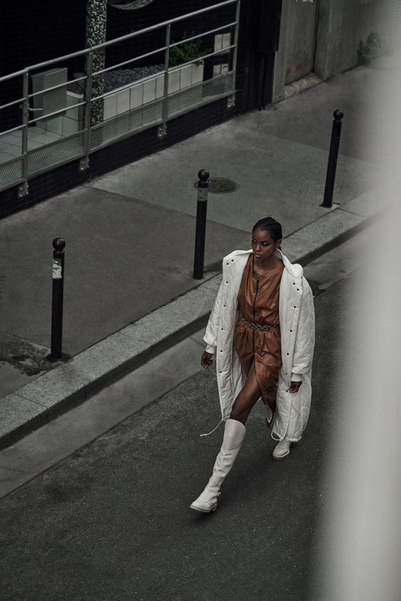 Fashionable woman in brown dress and white coat walks confidently down a city street in chic urban setting.