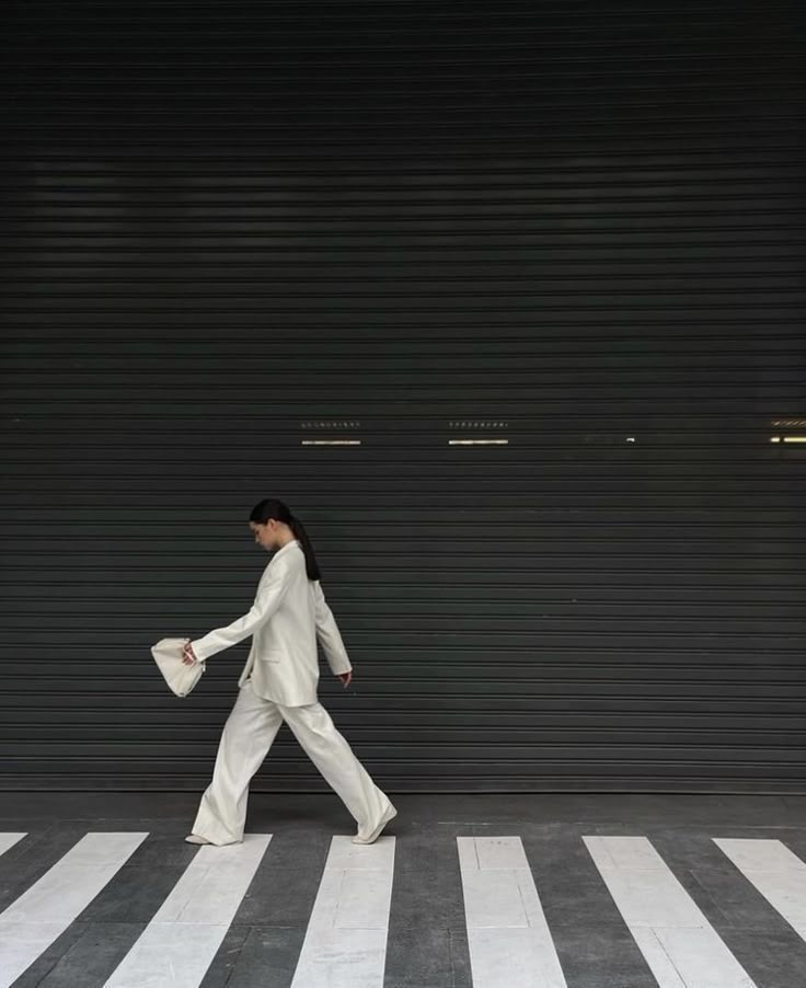 Person in a white suit walking on a zebra crossing against a dark background, holding a small bag.