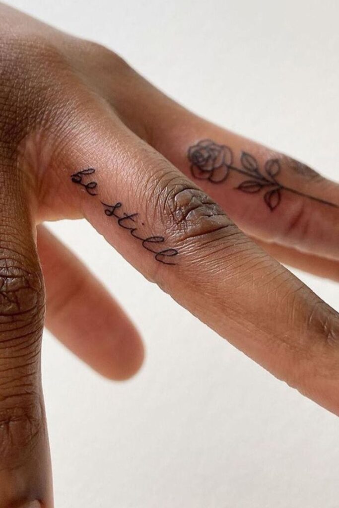 Close-up of finger tattoos with be still text and a small rose design. Minimalistic black ink art on skin.