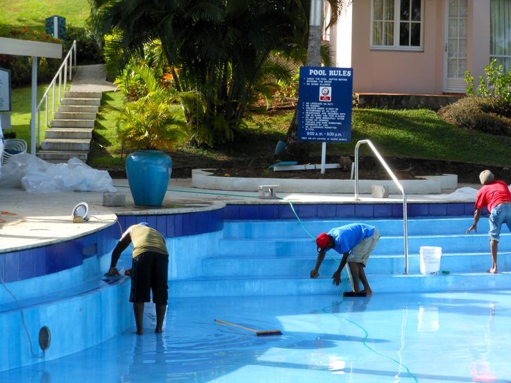 Workers cleaning an empty pool with tools and hose, near lush green garden and pool rules sign.