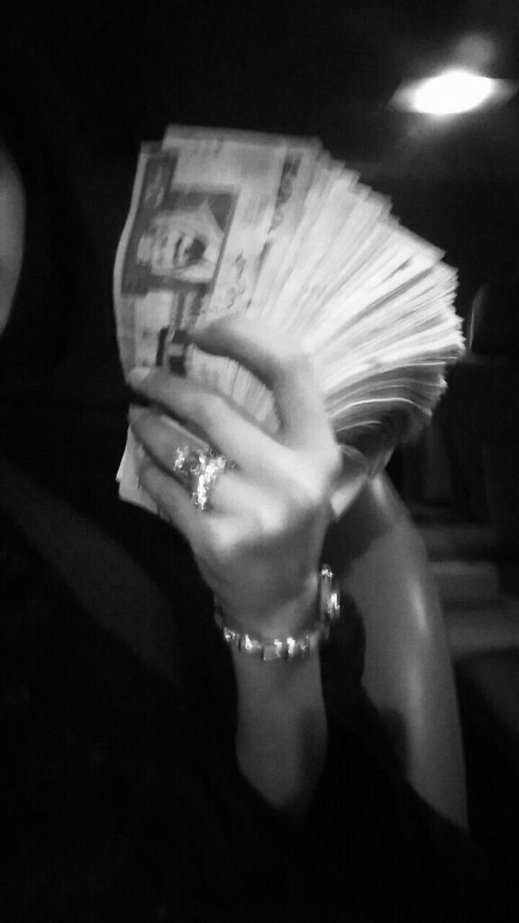 Person holding a large stack of currency notes with decorated hand in a dimly lit setting.