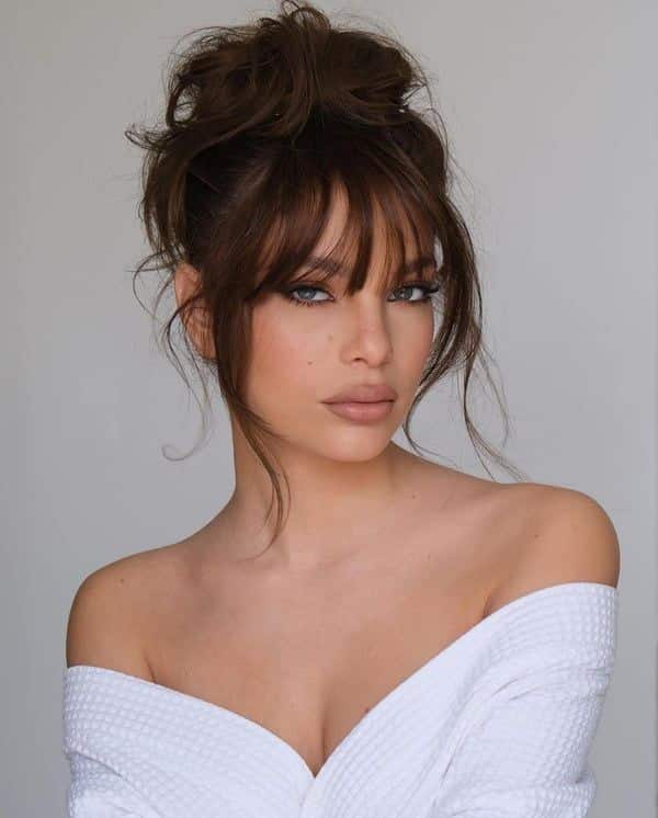 Woman with brown hair and bangs in a white robe, gazing confidently at the camera with a minimalist background.