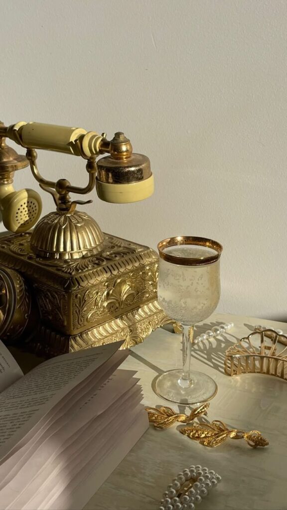 Vintage gold telephone with wine glass, pearls, book, and hair accessories in soft light. Elegant and timeless decor.