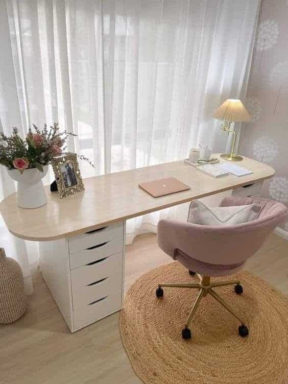 Elegant home office with a wooden desk, pink chair, laptop, and lamp, surrounded by soft, natural light through curtains.