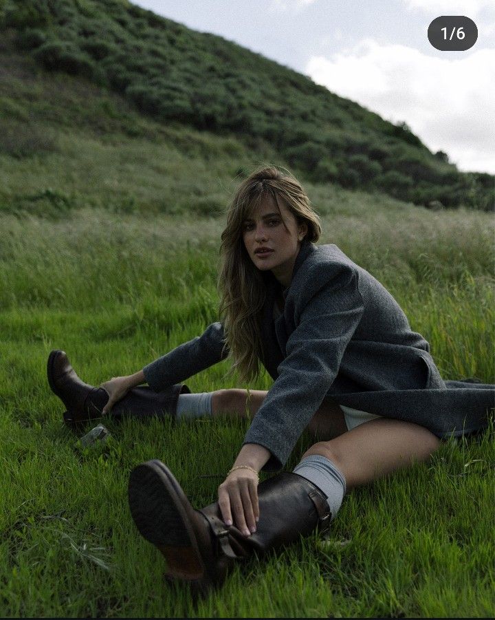 Person in a blazer and boots sitting on grass, with a scenic hillside backdrop.