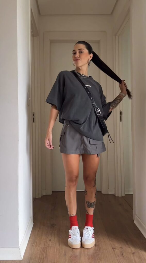 Fashionable woman in gray outfit with tattoos, red socks, and sneakers, standing confidently in a hallway.