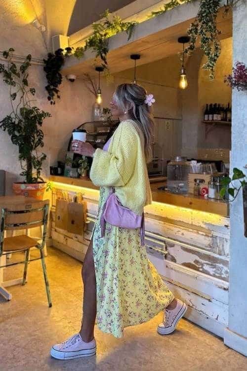 Woman in a floral dress and sneakers holding coffee walks through a cozy café with plants and warm lighting.