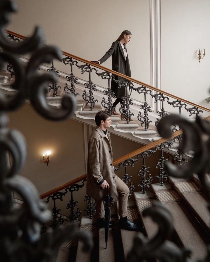 Elegant individuals descending ornate staircase with intricate wrought iron railings in a classic interior setting.