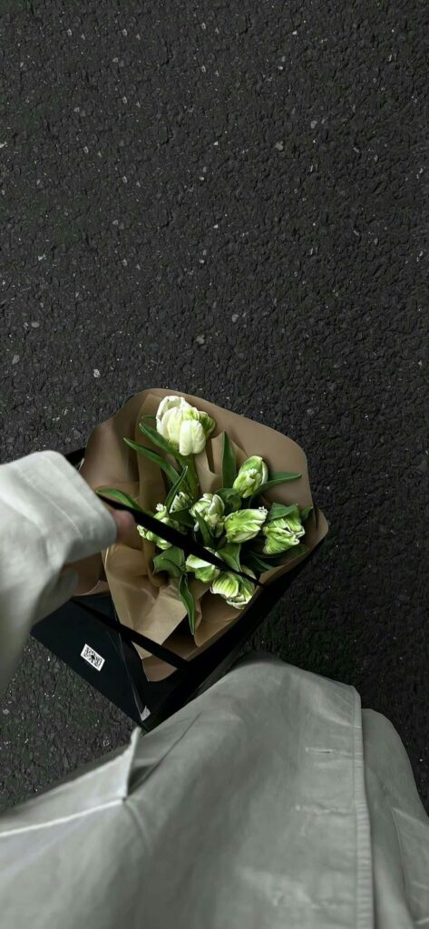 Person holding a bouquet of white and green flowers in a black paper bag on a textured gray surface.