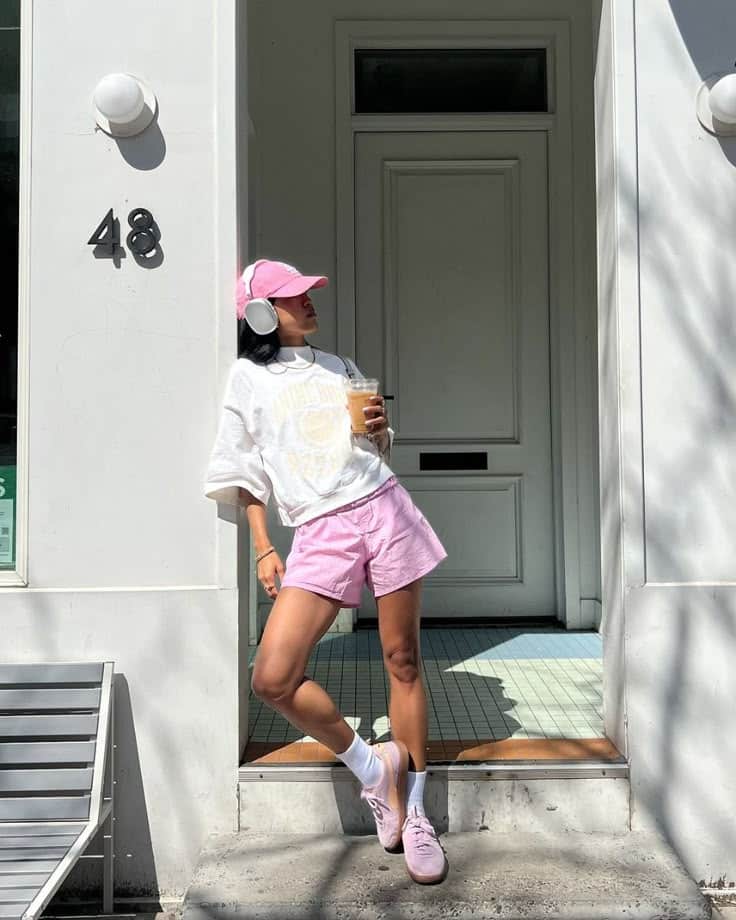 Person in pink shorts and cap holds iced coffee, leaning against building entrance with number 48.