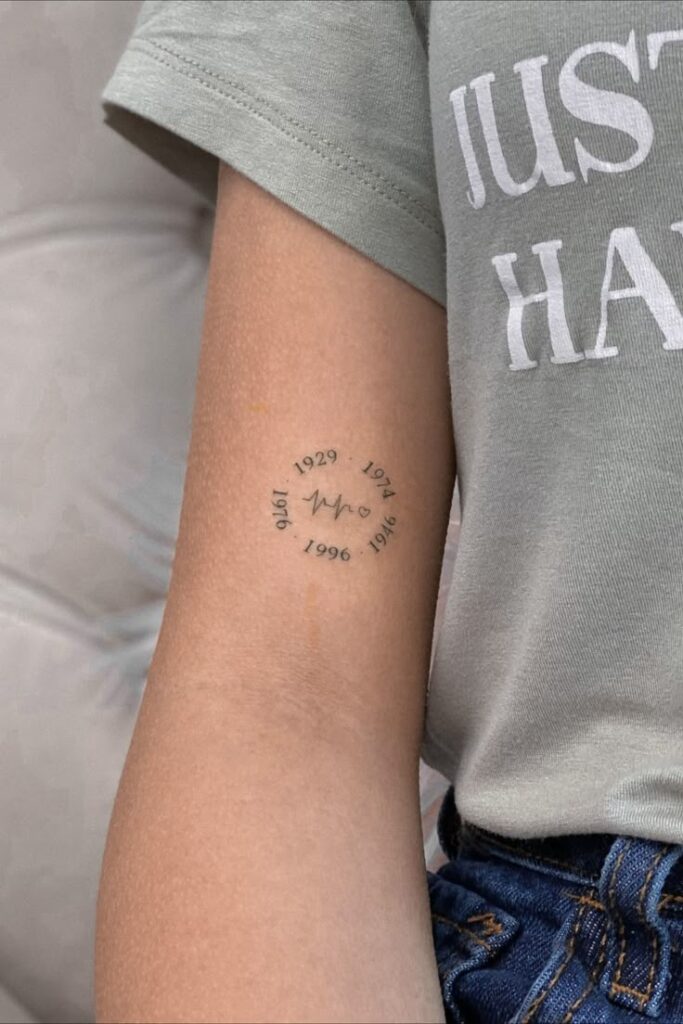 Arm tattoo of dates encircling heartbeat design, symbolizing personal milestones, on person wearing a green t-shirt.