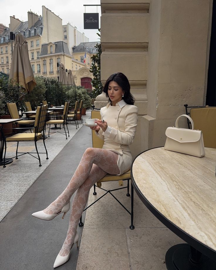 Woman enjoys coffee at a stylish Parisian cafe, wearing elegant white outfit with lace tights and heels.