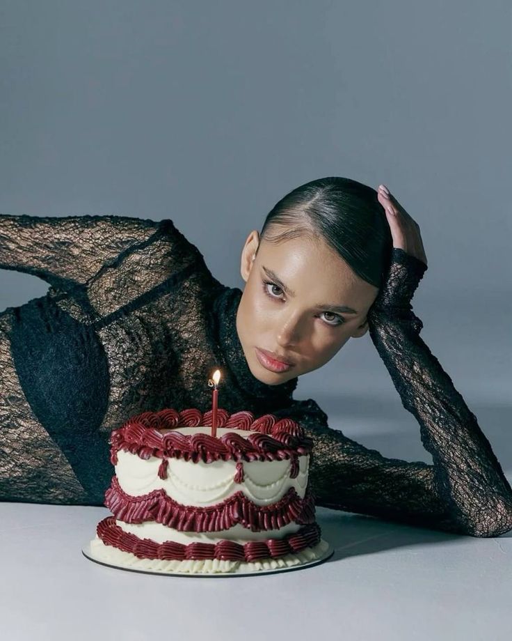 Person in black lace outfit posed with a decorative cake, candle lit, on a minimalist gray background.