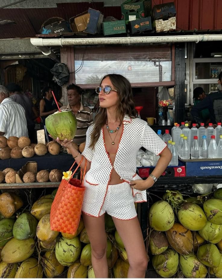 Woman in stylish beachwear with coconut at tropical market, colorful bag and sunglasses.