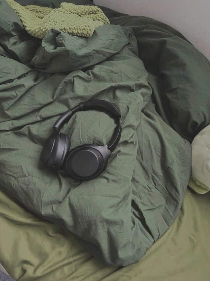 Black headphones on a crumpled green comforter with a knitted green throw blanket on a bed.