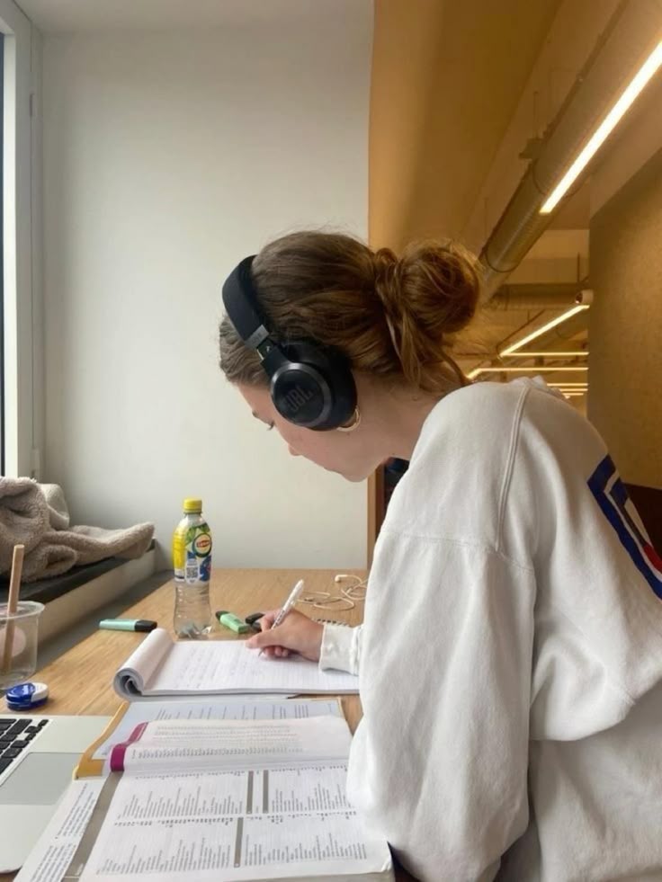 Person studying at a desk with headphones, writing notes and using a laptop, surrounded by books and a drink bottle.
