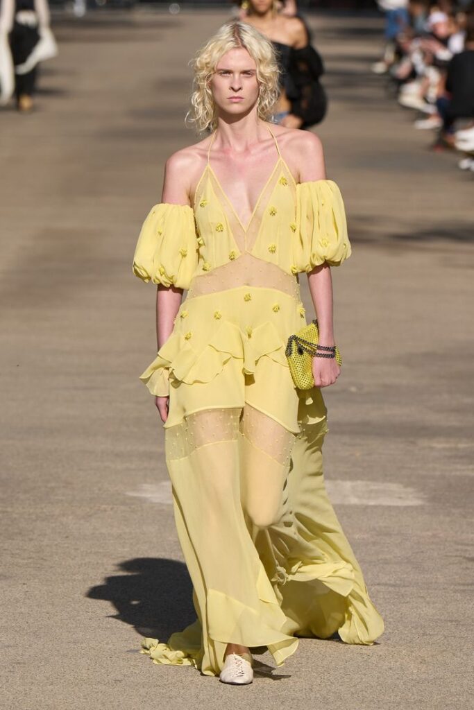Model walking runway in sheer yellow dress with puff sleeves, featuring floral accents, at a fashion show.