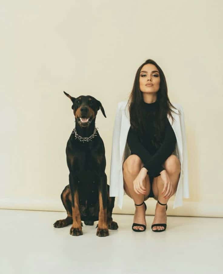 Woman in white jacket and black dress sitting next to a Doberman against a beige background.