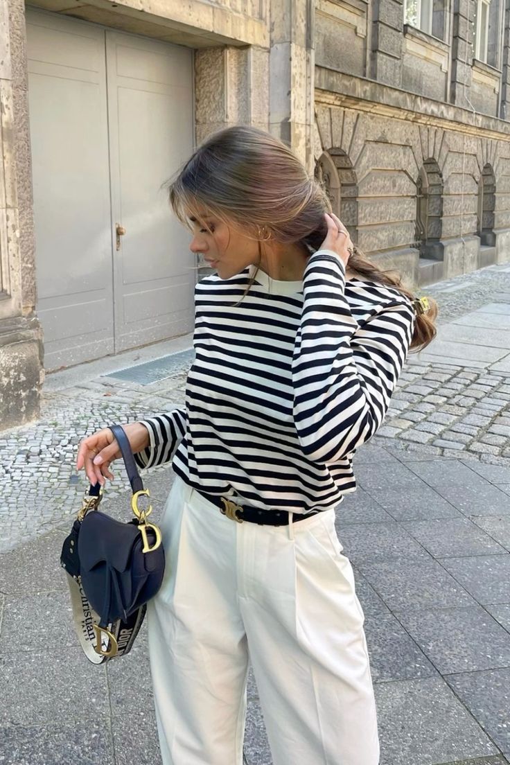 58bf411c-c1c5-4867-a1b8-aeb547cd005d Fashionable woman in striped shirt and white pants holding a stylish handbag, walking in an urban setting. | Sky Rye Design Fashionable woman in striped shirt and white pants holding a stylish handbag, walking in an urban setting.