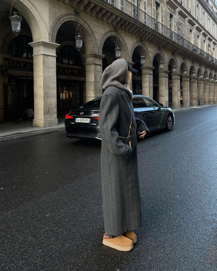 Person in long coat and beanie stands by street, elegant architecture backdrop, black car parked nearby.