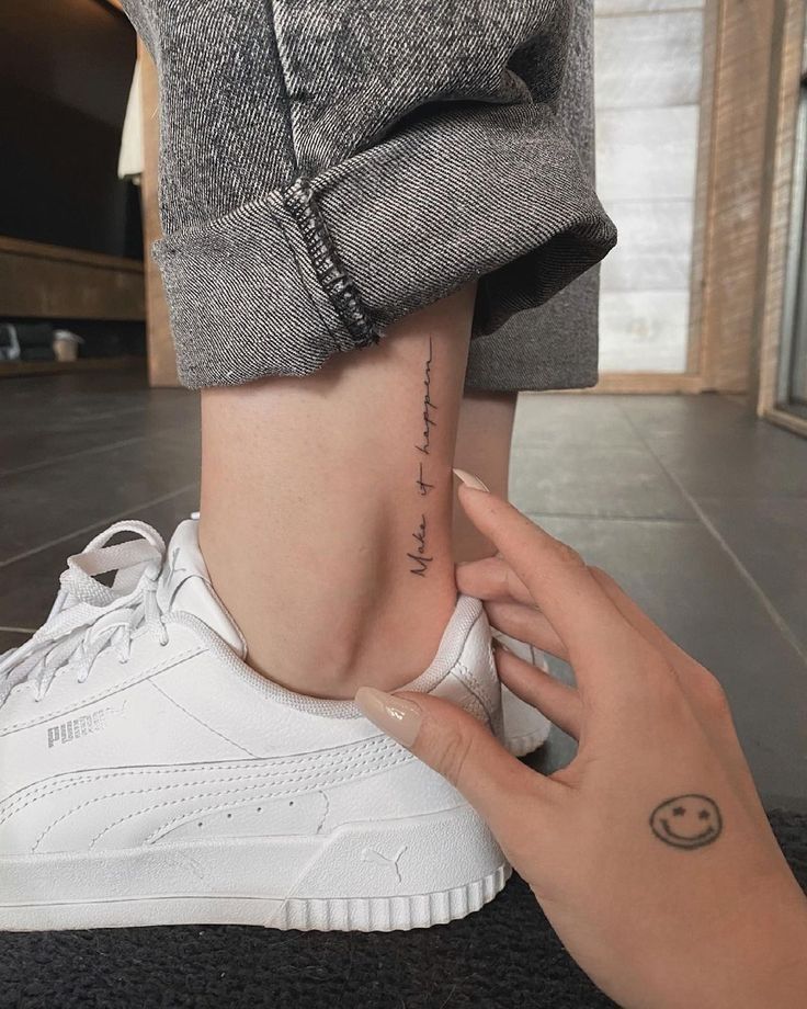 Close-up of ankle tattoo with script. Person wearing white sneakers and rolled-up jeans, hand touching tattooed area.
