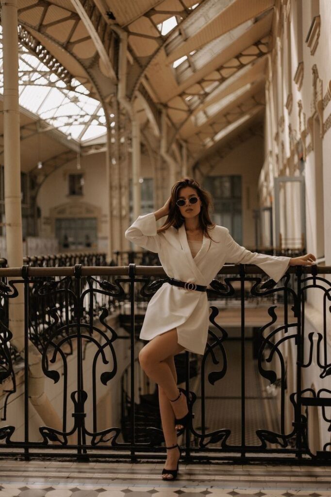 Fashionable woman in white blazer dress and sunglasses posing by vintage railing indoors.