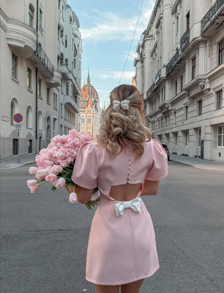 Woman in pink dress holding tulips, walking in a European city street with historic architecture in the background. Pink Aesthetic Wallpaper
