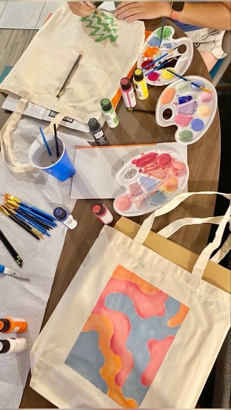 5d056c71-1297-40ec-81de-05bf3431367f Tote bags being hand-painted with colorful designs, surrounded by paints, brushes, and art supplies on a wooden table. | Sky Rye Design Tote bags being hand-painted with colorful designs, surrounded by paints, brushes, and art supplies on a wooden table.