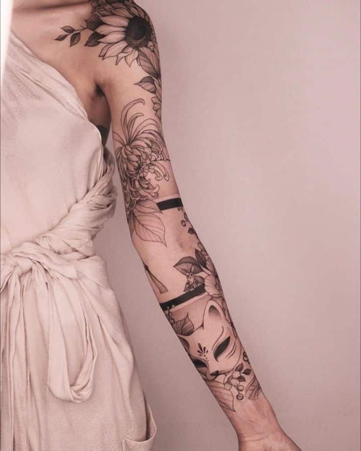 Floral and fox mask tattoo sleeve on woman's arm in beige dress, showcasing intricate linework.