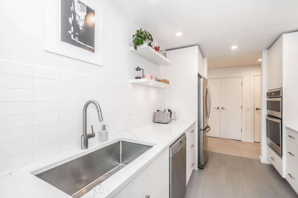 Modern white kitchen with stainless steel appliances, marble countertop, and open shelving. Minimalist design.