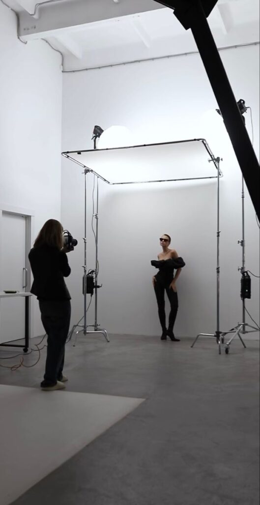 Photography studio session with a model posing under lights while photographer captures the scene.