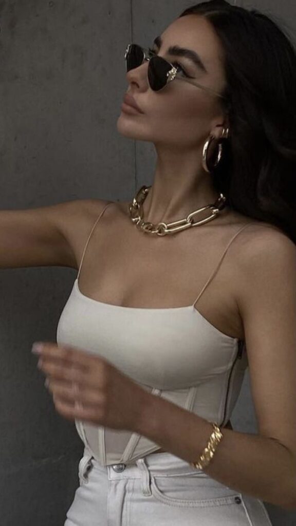 Woman in chic outfit with sunglasses and gold accessories, wearing a white top and jeans against a gray background.