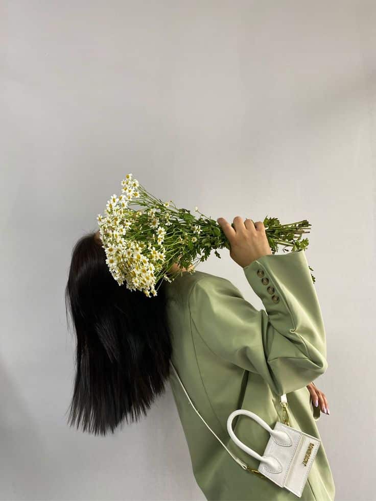 Woman in green blazer holding daisies, white mini handbag, hair flowing, against gray background. Fashion and nature blend.