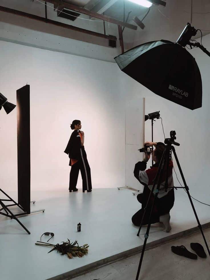 Fashion photoshoot in studio with model posing and photographer capturing on camera, surrounded by lights and equipment.