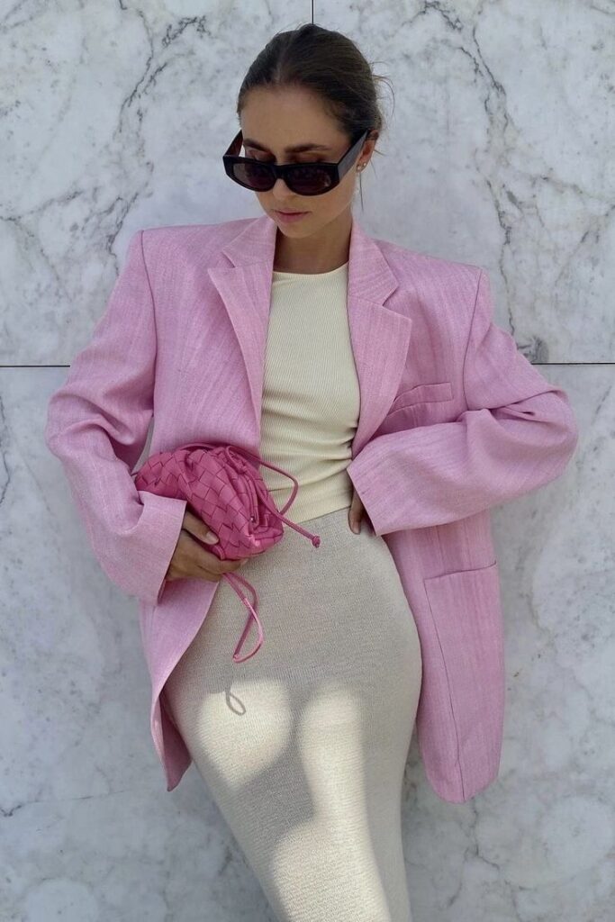 Stylish woman in oversized pink blazer and sunglasses, holding a pink bag against a marble wall. Fashionable look.