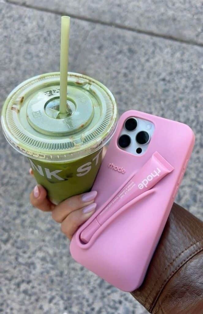 Hand holding matcha iced drink and pink phone with a pink case and lip balm, outdoors on pavement.