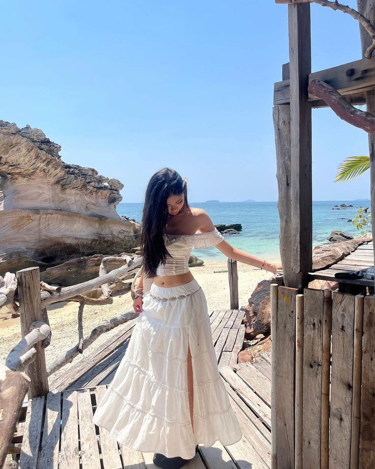 Woman in white dress on a sunny beach boardwalk, with blue sky and sea view in the background.