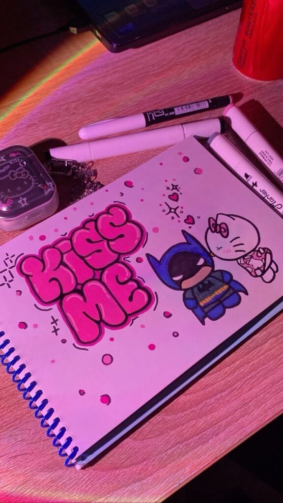 Notebook with cute doodles of a superhero and a character, alongside Kiss Me text, surrounded by markers.