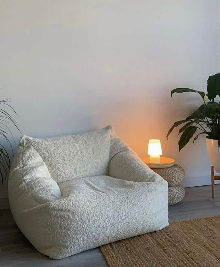 Cozy white armchair in a minimalist living room with a small lamp and potted plant on a woven side table.
