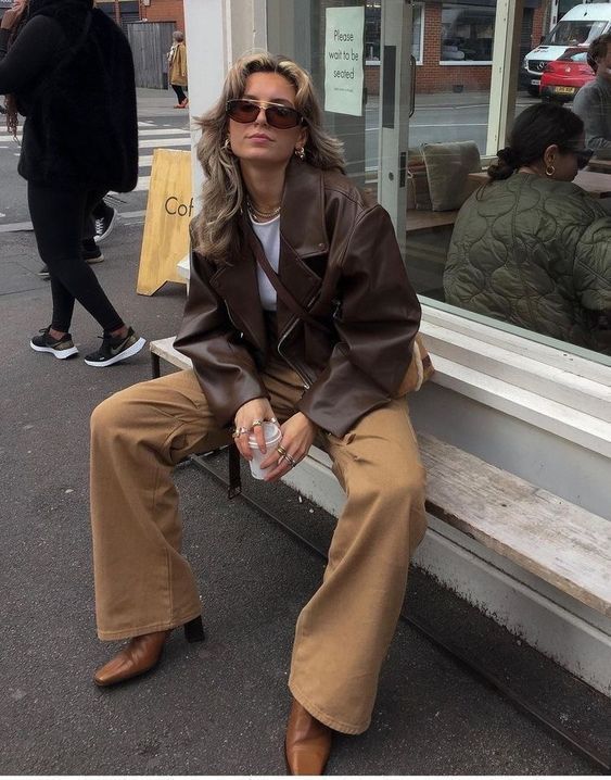 Fashionable woman in a brown leather jacket and tan pants sitting outside a café, holding a drink.