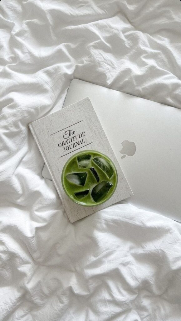 Gratitude journal and matcha drink on a laptop over white bedding, promoting mindfulness and productivity.