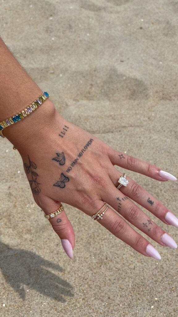 Hand with butterfly tattoos, colorful bracelet, rings, and pink nails against sandy background.