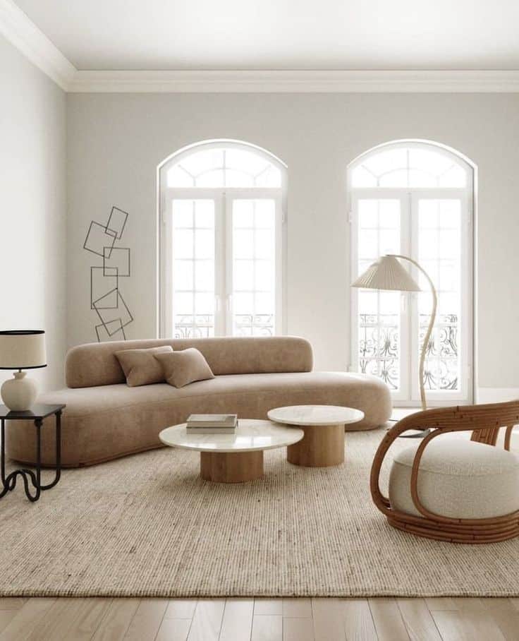 Modern living room with beige sofa, round tables, wicker chair, and large windows. Elegant and minimalist decor.