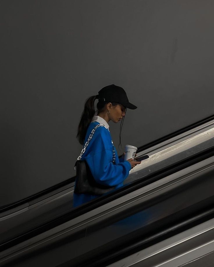 Person in blue sweatshirt on escalator, holding coffee and smartphone, wearing cap and black bag against gray background.