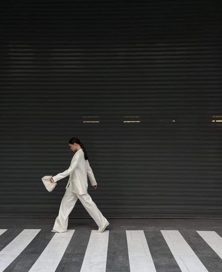 Woman in white suit walking on a zebra crossing, holding a handbag against a dark shutter background.